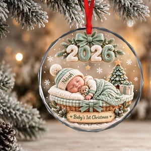 Könnte beinhalten: Ein runder, transparenter Acryl-Weihnachtsanhänger mit einem schlafenden Baby im Korb, das eine Strickmütze mit der Aufschrift Noah trägt. Das Design zeigt die Zahl 2026, einen kleinen Weihnachtsbaum, einen Teddybären und Baby's 1st Christmas.