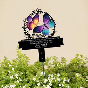 Könnte beinhalten: Ein schwarzer Metall-Gartenstecker mit einem Schmetterlingsdesign mit lila, gelben und türkisfarbenen Flügeln, umgeben von einem floralen Kranz. Der Stecker trägt den Text "Those we love don't go away... Amy Smith 12/01/1931-11/16/2025."