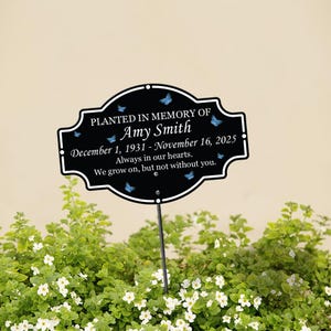 Könnte beinhalten: Ein schwarzer Gedenk-Gartenstecker mit weißem Text und blauen Schmetterlingsakzenten. Der Text lautet "Gepflanzt in Erinnerung an Amy Smith" mit Daten und einer herzlichen Botschaft. Der Stecker befindet sich in einem Beet mit grünen Pflanzen und weißen Blüten.