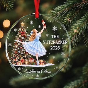 Könnte beinhalten: Ein runder, klarer Glas-Weihnachtsbaumschmuck mit einer gedruckten Illustration einer Ballerina im blauen Kleid, die einen Nussknacker hält. Das Design zeigt einen geschmückten Weihnachtsbaum, Geschenke, Schneeflocken sowie den Text THE NUTCRACKER 2026 und Sophia as Clara.