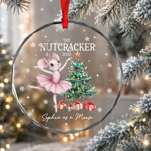 May include: A round, clear glass Christmas ornament with a red ribbon hanger. It features a white mouse in a pink tutu, a decorated Christmas tree with gifts, and the text: THE NUTCRACKER 2026, Sophia as a Mouse, surrounded by white snowflakes.