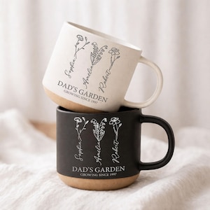 Könnte beinhalten: Zwei Keramikbecher, einer weiß und einer schwarz, übereinander gestapelt. Die Becher haben einen beigefarbenen Boden und schwarze Henkel. Jeder Becher zeigt Blumenillustrationen und den Text "DAD'S GARDEN GROWING SINCE 1997" mit den Namen Sophia, Amelia und Robert.