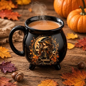 Puede incluir: Una taza de cerámica negra con forma de caldero que presenta un gráfico de una bruja sosteniendo una linterna bajo una luna creciente, rodeada de hojas de otoño. El nombre Sophia está impreso en letras doradas bajo la ilustración.