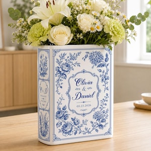 Könnte beinhalten: Eine weiße, buchförmige Keramikvase mit blauen Blumenmustern und dekorativen Rändern. Auf der Vorderseite steht der Text Olivia & Daniel 05.23.2026, an der Seite C + D ENGAGED 05.23.2026. Die Vase ist mit weißen Lilien, Rosen und Grünpflanzen gefüllt.