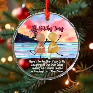 Könnte beinhalten: Ein klarer, runder Weihnachtsschmuck mit einer Strandszene und dem Text "The Bitches Times". Zwei Strichmännchen, beschriftet mit "Amelia" und "Jessica", sind abgebildet. Der Schmuck enthält auch den Text "Here's To Another Year Of Us..."