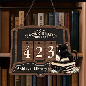 Könnte beinhalten: Holzschild mit dem Text "BOOK READ THIS YEAR" und der Zahl "423". Eine schwarze Katze sitzt auf einem Stapel Bücher, umgeben von Blumen. Das Schild trägt auch die Aufschrift "Ashley's Library".