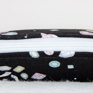 Puede incluir: Una bolsa negra con cremallera blanca. La bolsa est&aacute; cubierta con un patr&oacute;n de piedras preciosas de colores.