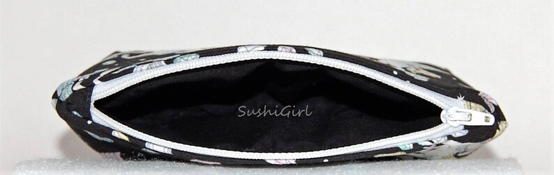 Puede incluir: Bolsa negra con cremallera blanca y dise&ntilde;o estampado en blanco y negro. El interior de la bolsa es negro y tiene el texto "SushiGirl" impreso en blanco.