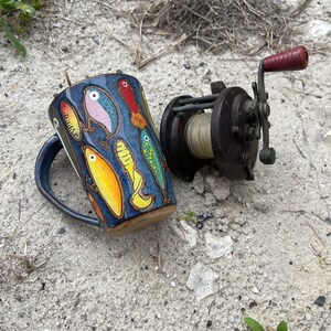 May include: A blue ceramic mug with a colorful design of fishing lures. The mug is next to a vintage fishing reel with a red handle.
