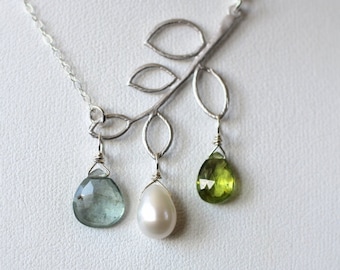 May include: A silver necklace featuring a leaf design with three dangling pendants. The pendants include a pear-shaped blue-green gemstone, a white pearl, and a green gemstone. The necklace is displayed on a white surface.