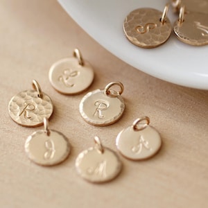 May include: Gold-toned metal charms with different letters engraved on them. The charms are round and have a hammered texture.