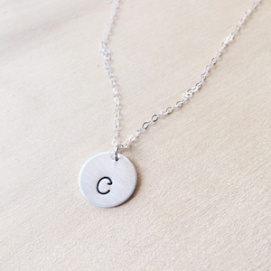 May include: A silver-colored necklace with a circular pendant. The pendant has the letter "C" in black. The necklace chain is delicate and hangs on a light-colored surface.