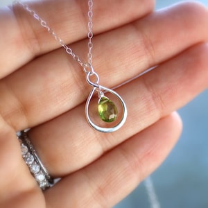 May include: A delicate silver necklace featuring a teardrop-shaped pendant. The pendant holds a vibrant, pear-cut peridot gemstone. The necklace is displayed against a soft, blurred background, highlighting the jewelry's elegant design.