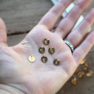 May include: A hand holding several small gold charms with letters on them. The charms are round and have a hole for a chain or string.