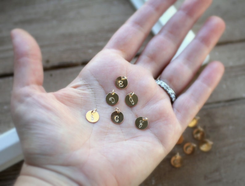May include: A hand holding several small gold charms with letters on them. The charms are round and have a hole for a chain or string.