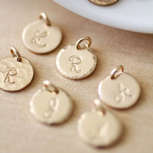 May include: A collection of gold-toned metal charms with different letters engraved on them. The charms are round and have a hammered texture.