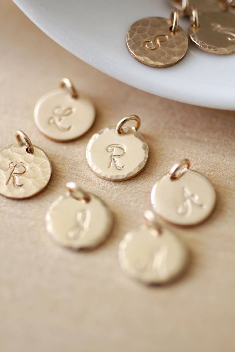 May include: A collection of gold-toned metal charms with different letters engraved on them. The charms are round and have a hammered texture.