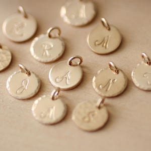 May include: A collection of 10 gold-toned metal charms with different letters engraved on them. The charms are round and have a textured surface.
