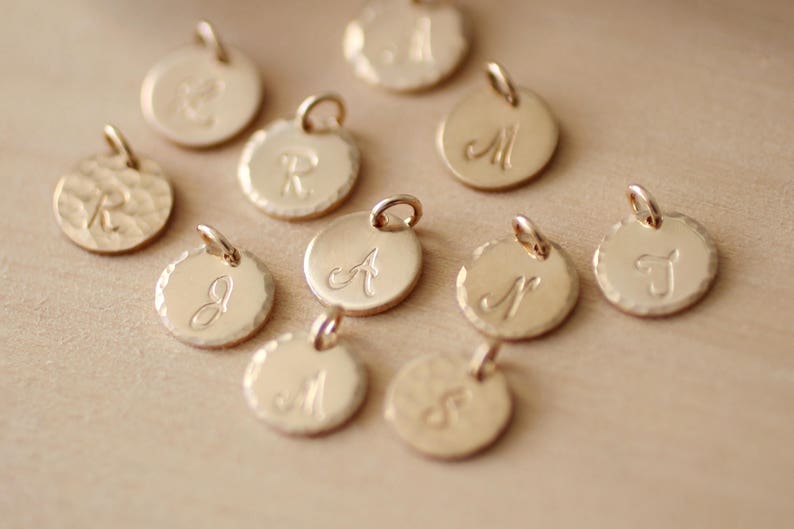 May include: A collection of 10 gold-toned metal charms with different letters engraved on them. The charms are round and have a textured surface.