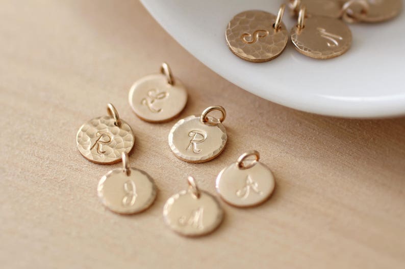 May include: Gold-toned metal charms with different letters engraved on them. The charms are round and have a hammered texture.