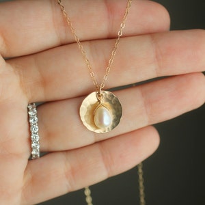 May include: A gold necklace with a hammered disc pendant and a single pearl. The necklace is on a hand wearing a diamond ring.