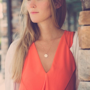 May include: A woman with long blonde hair wearing a bright orange sleeveless dress with a gold necklace and a round pendant. She is wearing a white cardigan and looking off to the side.