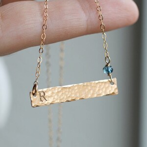 May include: Gold necklace with a hammered bar pendant and a small blue gemstone. The bar has the letter "R" engraved on it.