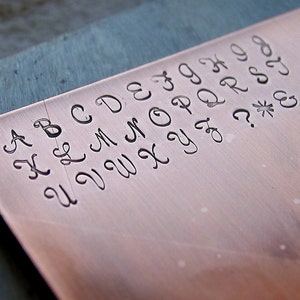 May include: A close-up of a copper plate with the alphabet engraved in a cursive font. The letters are A, B, C, D, E, F, G, H, I, J, K, L, M, N, O, P, Q, R, S, T, U, V, W, X, Y, Z, and punctuation marks including a question mark, asterisk, and ampersand.