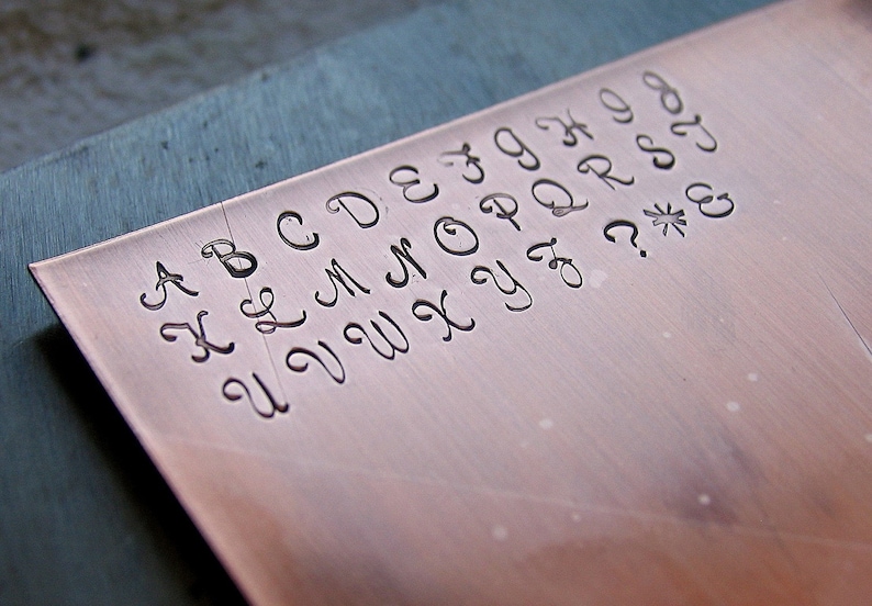 May include: A close-up of a copper plate with the alphabet engraved in a cursive font. The letters are A, B, C, D, E, F, G, H, I, J, K, L, M, N, O, P, Q, R, S, T, U, V, W, X, Y, Z, and punctuation marks including a question mark, asterisk, and ampersand.