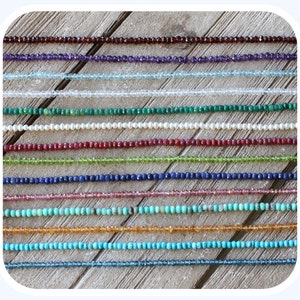 May include: A collection of 18 strands of gemstone beads, each strand is a different color and type of gemstone. The beads are arranged in rows on a wooden surface.