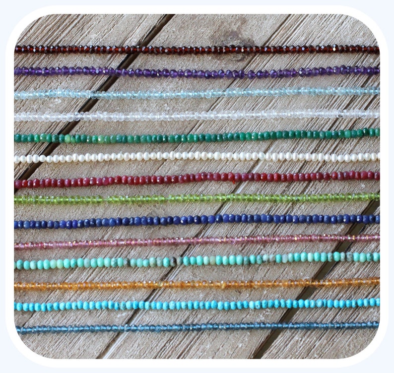 May include: A collection of 18 strands of gemstone beads, each strand is a different color and type of gemstone. The beads are arranged in rows on a wooden surface.