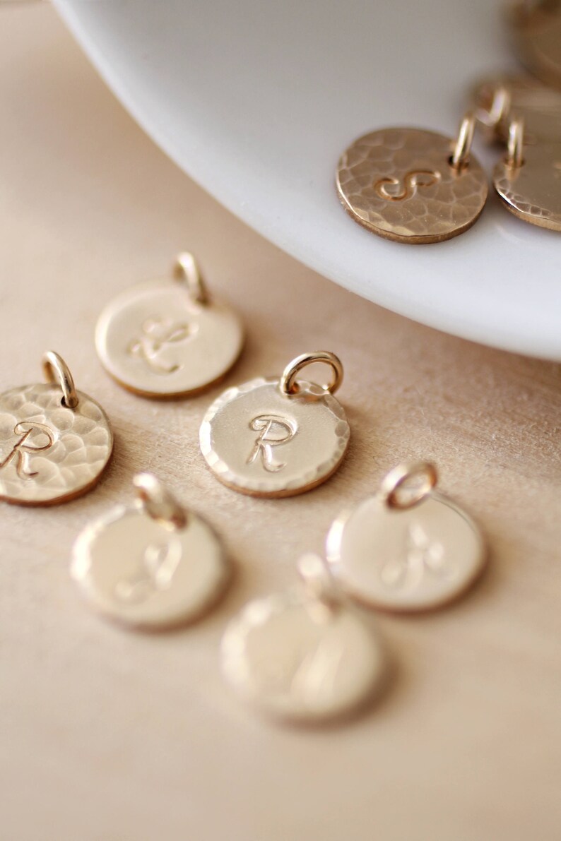 May include: A collection of gold-toned metal charms with different letters engraved on them. The charms are round and have a hammered texture.