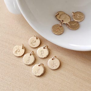 May include: A collection of gold-toned metal charms with different letters engraved on them. The charms are round and have a hammered texture.