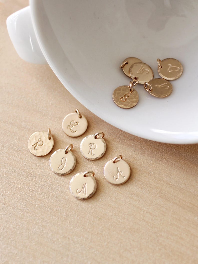 May include: A collection of gold-toned metal charms with different letters engraved on them. The charms are round and have a hammered texture.