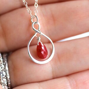 May include: A silver infinity necklace with a red gemstone pendant. The pendant is a teardrop shape and the gemstone is faceted.