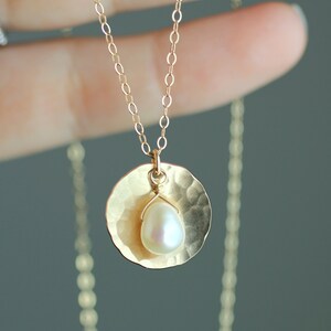 May include: A gold necklace with a hammered disc pendant and a single pearl. The necklace is on a model's hand.