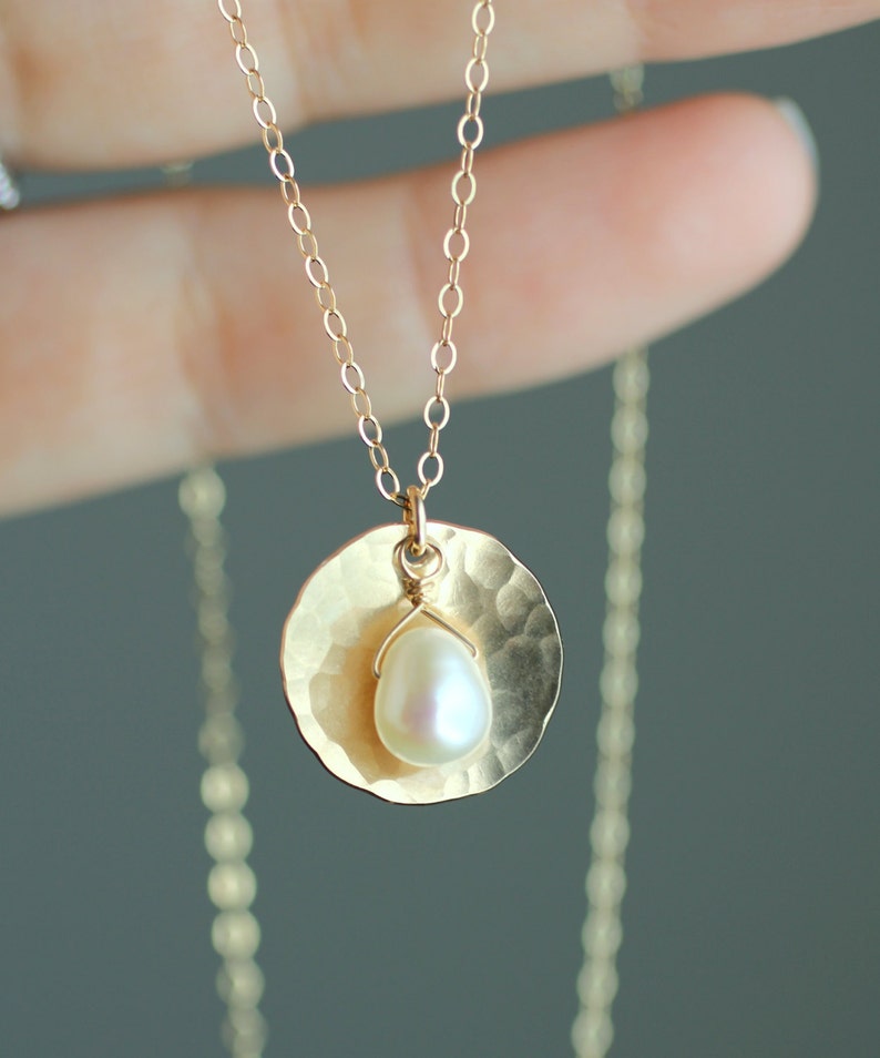 May include: A gold necklace with a hammered disc pendant and a single pearl. The necklace is on a model's hand.