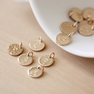 May include: A collection of gold-toned metal charms with different letters engraved on them. The charms are round and have a hammered texture.