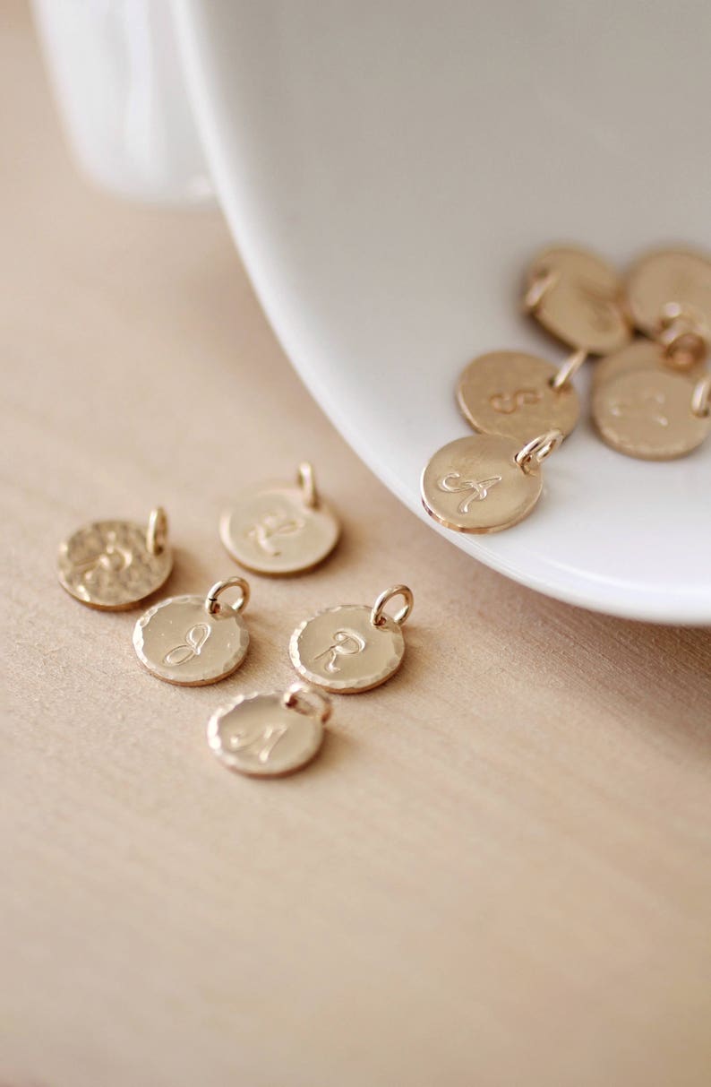 May include: A collection of gold-toned metal charms with different letters engraved on them. The charms are round and have a hammered texture.