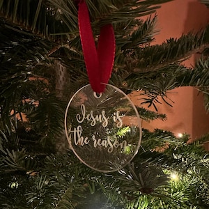 Könnte beinhalten: Ein klarer, runder Weihnachtsschmuck mit dem Text "Jesus is the reason" in Schreibschrift. Der Schmuck hängt an einem roten Band vor dem Hintergrund eines Weihnachtsbaums.