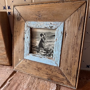 Op de afbeelding: Een vierkante fotolijst gemaakt van gerecycled hout met een natuurlijke bruine afwerking en een verweerde lichtblauwe binnenrand. De lijst bevat een zwart-witfoto van een figuur in een uitgestrekt, open landschap.