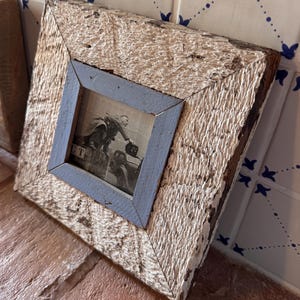 Op de afbeelding: Vierkante houten fotolijst met een verweerde, getextureerde afwerking. De buitenste lijst is lichtbeige met witte verf en de binnenste lijst is blauw. De zwart-witfoto toont een persoon op een auto.