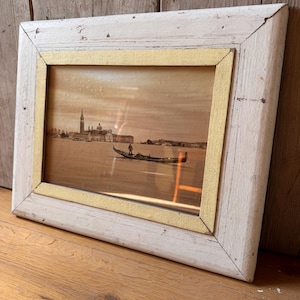 May include: An antique framed print of a Venetian scene. The print features a gondola on the water with buildings in the background. The frame is white with a gold inner border, showing signs of wear. The artwork is displayed on a wooden surface.