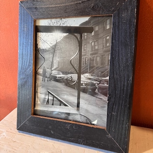 Puede incluir: Fotografía enmarcada en negro de una escena de calle urbana en blanco y negro. El marco es de madera oscura con veta visible. La fotografía representa coches aparcados, edificios y personas en una acera.