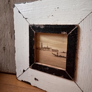 Op de afbeelding: Een rechthoekige, verweerde wit en zwart omlijste foto. De lijst heeft een getextureerd, verweerd uiterlijk. De foto binnenin toont een gondel op het water met gebouwen op de achtergrond. De lijst rust op een houten oppervlak.