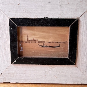 May include: An antique framed print depicting a Venetian scene. The frame is distressed white with a black inner border. The sepia-toned print shows a gondola on the water with buildings in the background, including a tall bell tower.