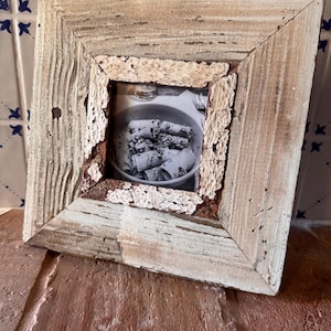 Op de afbeelding: Een vierkante fotolijst gemaakt van verweerd, lichtgekleurd hout met een verweerde witte afwerking. De binnenrand heeft een ruwe, getextureerde, witgeschilderde rand. Binnenin is een zwart-witfoto van pasta in een kom te zien.