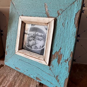 Op de afbeelding: Vierkante, verweerde fotolijst met een turquoise afwerking en een wit binnenframe. De lijst heeft een rustieke, verweerde uitstraling met zichtbare houtnerf en afbladderende verf, wat hem een vintage esthetiek geeft. De lijst bevat een zwart-wit foto.