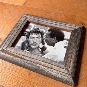 May include: A square, wooden picture frame with a black and white photograph. The photograph features two men, one with a mustache, in a close embrace. The frame has a weathered, gray finish and is resting on a wooden surface.