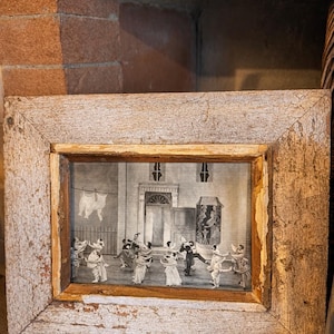 Puede incluir: Una impresión en blanco y negro enmarcada que representa una escena de baile con figuras bailando. El marco es de madera clara envejecida. La impresión muestra una ilustración detallada de una reunión social, con elementos arquitectónicos en el fondo.